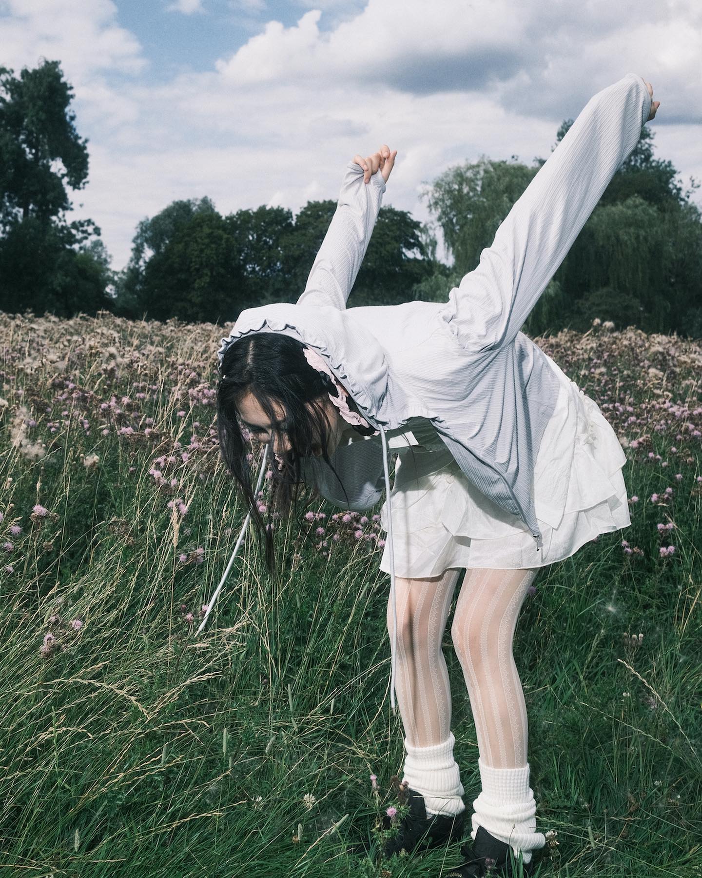 a young woman, possibly a model, playfully bends over in a field of wildflowers. she is wearing a light grey hooded jacket, a white dress, white striped tights, and black shoes. the setting appears to be a summer meadow on a partly cloudy day.  the overall mood of the image is ethereal and dreamy, with a touch of whimsy. the photo likely belongs to a fashion or artistic photoshoot.