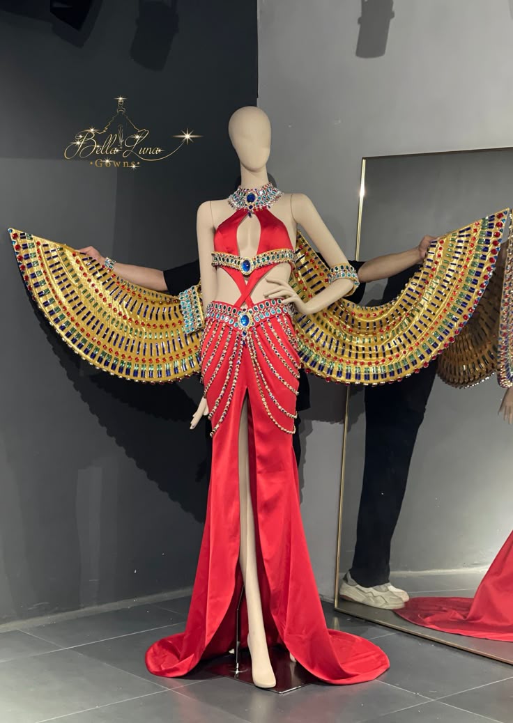a mannequin is dressed in a striking red, egyptianstyle gown with golden wings, adorned with jewels and beads. the dress features a high slit and cutout details, displayed against a dark gray wall and a partially visible mirror. a person stands beside the mannequin holding the wings in place.