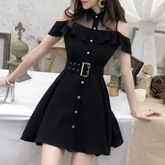 a young woman with long black hair is wearing a black short coldshoulder dress with a ruffled neckline. the dress is buttoned up the front and has a belt at the waist. she is standing in a room with white walls and some decor. the dress is cinched at the waist by a black belt with silver embellishments. the dress is styled with a delicate silver necklace. she is wearing light makeup and her nails are painted black. she is posing with her arm on a table and looking at the camera.  she is in front of a white background. she is wearing minimal jewelry.