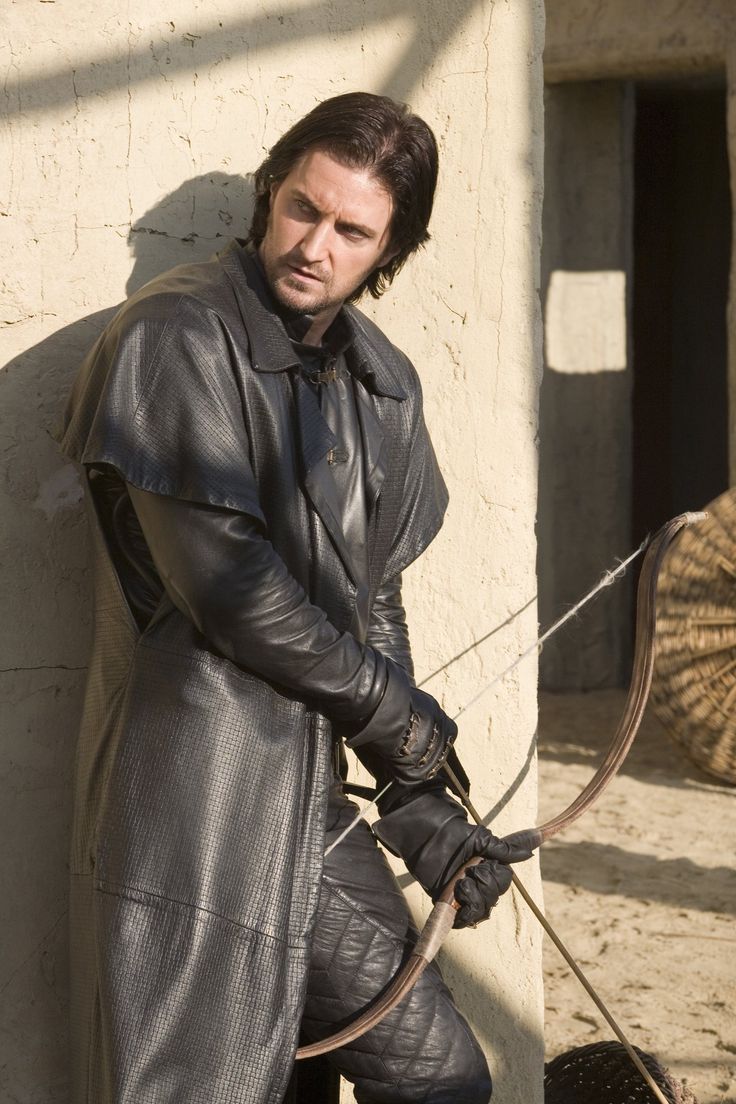 a man in a black leather coat stands against a wall, holding a bow and arrow. he has long dark hair and a serious expression. the scene appears to be set outdoors in a sunny location.