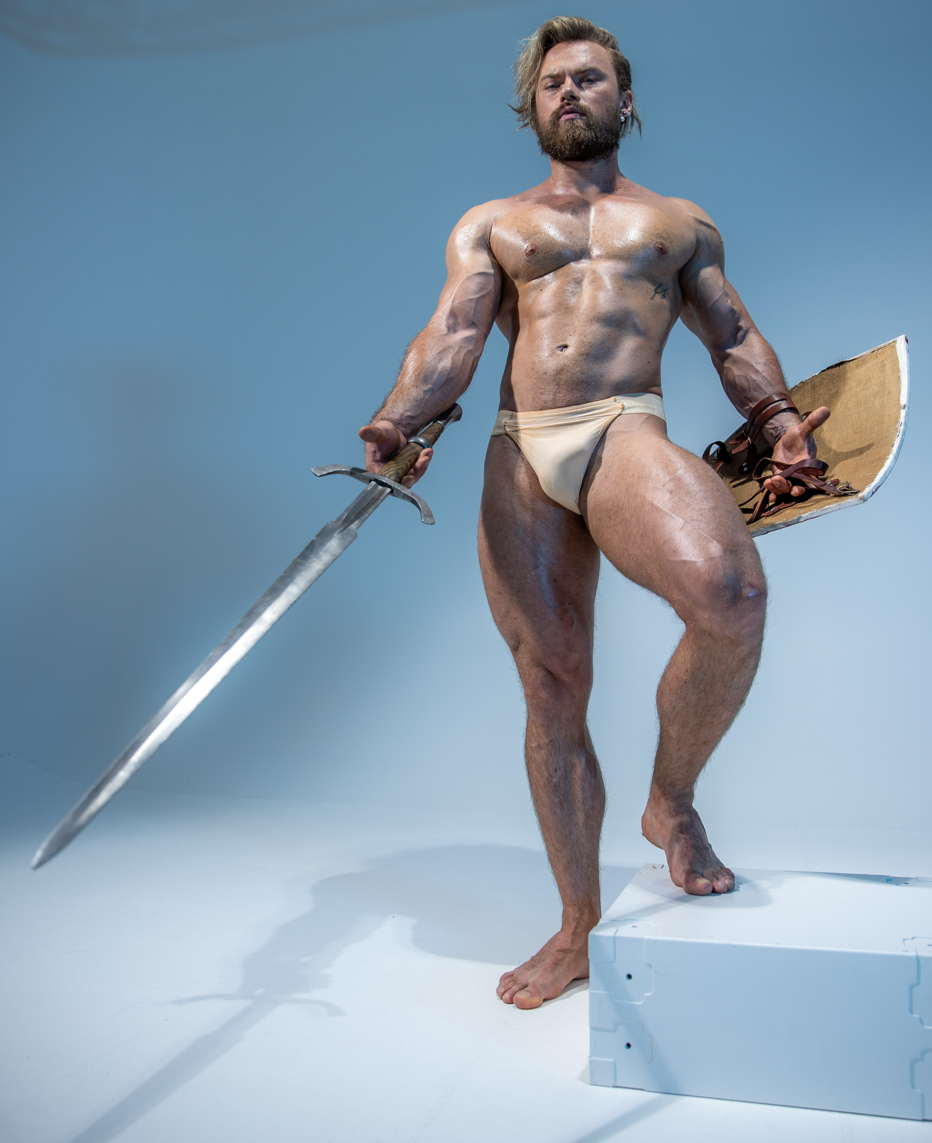 a muscular man with long blond hair and beard stands in a confident pose against a light blue background. he is wearing a loincloth and holds a sword in his right hand and a round shield in his left. the image evokes a sense of strength, power, and warrior spirit, reminiscent of mythological figures like vikings. the soft lighting highlights his physique and creates a dramatic atmosphere.