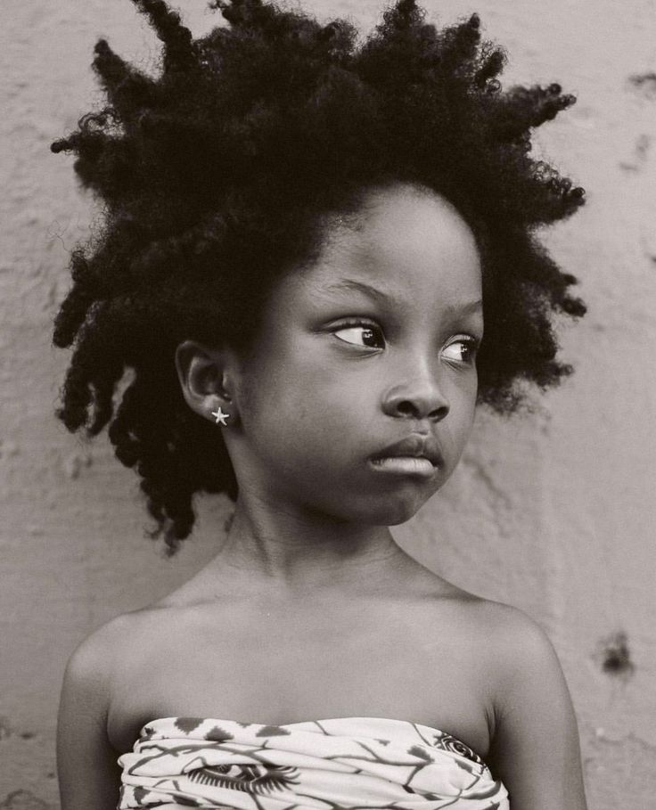 the image is a black and white closeup portrait of a young black girl with a large, voluminous afro. she's wearing a strapless top with a patterned design and a small starshaped earring. her expression is serious and somewhat pensive, and she is looking slightly away from the camera. the background is a textured wall, suggesting an outdoor setting. the overall mood of the image is contemplative and evokes a sense of strength and quiet dignity.  the photograph focuses on the subject's striking features and impressive hair, highlighting the texture and beauty of her natural hair.