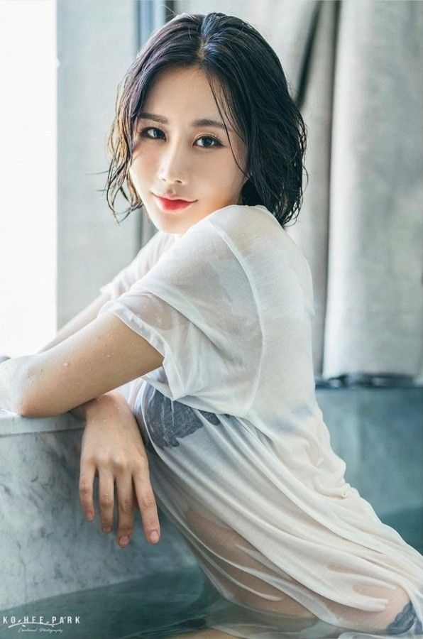 a closeup portrait of a young, attractive asian woman with short, wet dark hair. she is wearing a sheer, white tshirt that is clinging to her body due to the water. she is sitting in a bathtub with a marble edge, looking directly at the camera. the overall mood is sensual and serene. the photo is professionally taken and welllit, showcasing the model's beauty and the luxurious setting. the watermark "ko hee park" is visible in the bottom left corner.