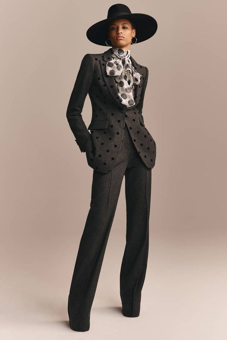 a woman in a black suit with a white polka dot blouse, a black fedora hat, and black heels is standing in a studio setting. the model is looking at the camera. the background is a light brown color.