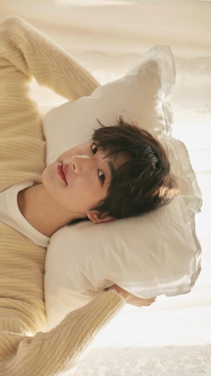 a young  asian man is relaxing on a  bed, his head resting comfortably on a fluffy white pillow. he's wearing a cozy, creamcolored sweater and looking directly at the camera with a relaxed expression. the lighting is soft and natural,  creating a peaceful and comfortable atmosphere.