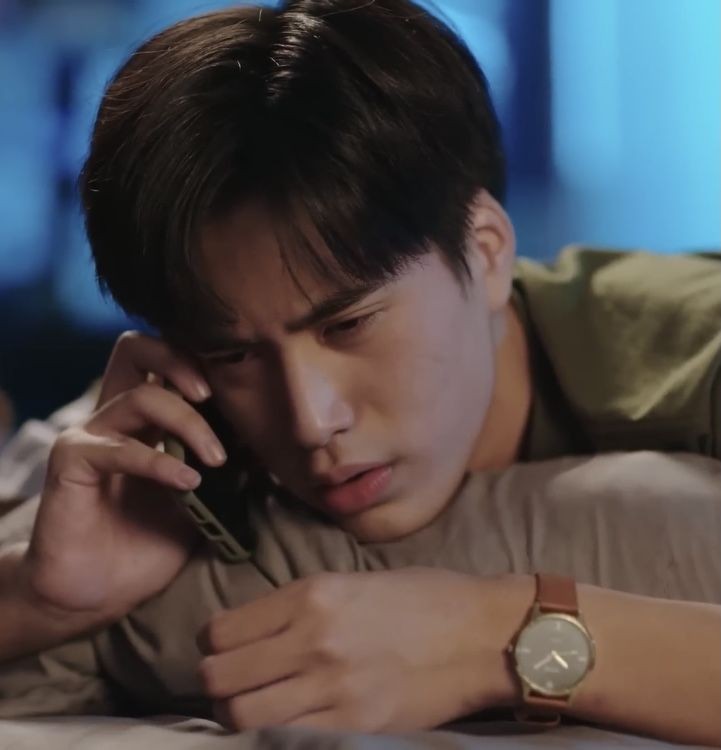 a young man lies on his stomach on a bed, holding a phone to his ear with a serious and concerned expression.  the lighting is dim and creates a sense of worry.