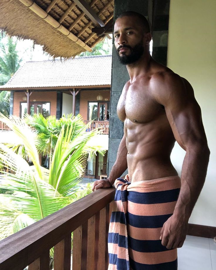a fit, muscular man stands on a balcony, wrapped in a striped towel. he is looking out at the view, with a palm tree in the foreground. the man is shirtless, showing off his defined six pack abs. the balcony is made of wood and has a railing, which is partially in focus. the man appears to be on vacation, in an exotic location. the background shows a tropical resort building with greenery and architecture. the image is welllit and the man is looking directly at the camera.