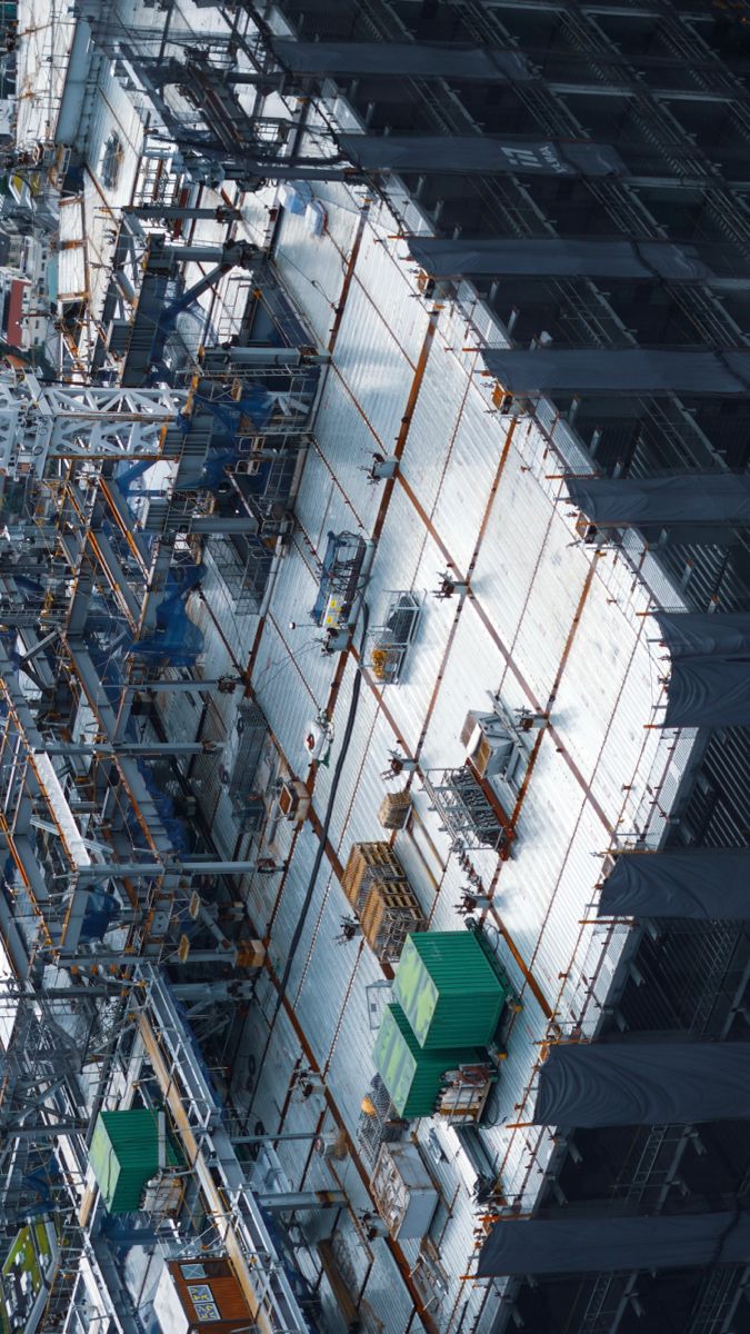 an overhead view of a large industrial construction site. a metal structure with scaffolding and a roof comprised of panels in a grid pattern is seen. containers and crates are scattered across the area, and various types of machinery are present.