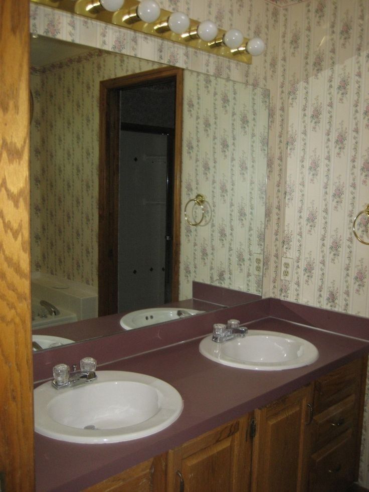a bathroom vanity with two sinks, a mirror, and a cabinet. the vanity top is a dark maroon color. the countertop has two sinks with chrome faucets. the mirror is over the sinks and shows a partial view of a bathroom with a toilet, a towel rack, and a tiled floor. the vanity is made of wood and has cabinets below the sink and on the right side of the vanity. the walls are covered in floral wallpaper.