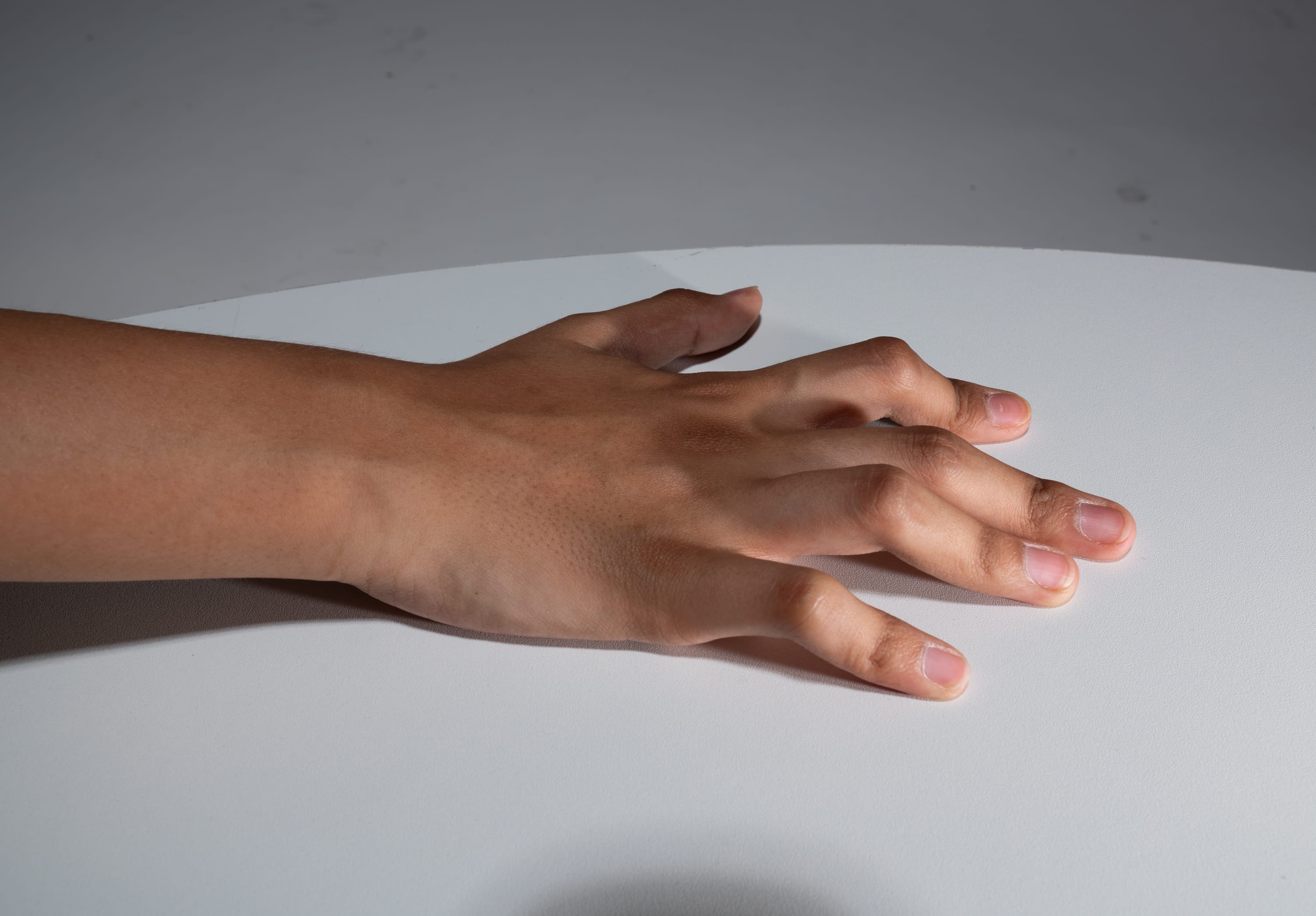 the image shows a hand resting on a white table. the hand is positioned with the palm facing down, and all five fingers are extended. the skin tone is light brown, and the image is illuminated with studio lighting.