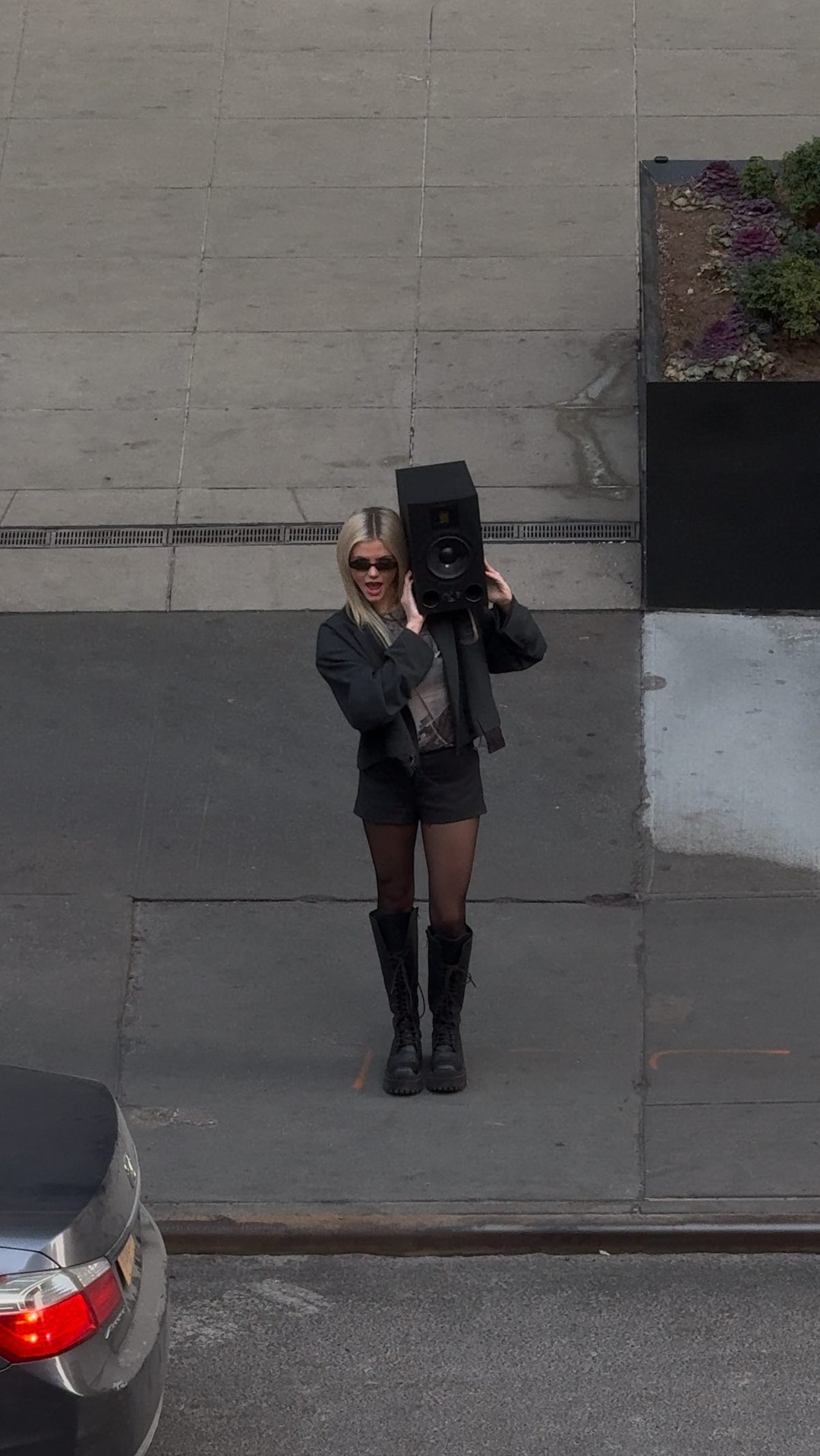 a woman with blonde hair, wearing a black jacket, black shorts, and black boots, stands on a pavement. she's holding a black speaker on her shoulder. she's also wearing sunglasses. a car is parked in the bottom left corner of the image. the background is an urban setting with pavement and a planter.