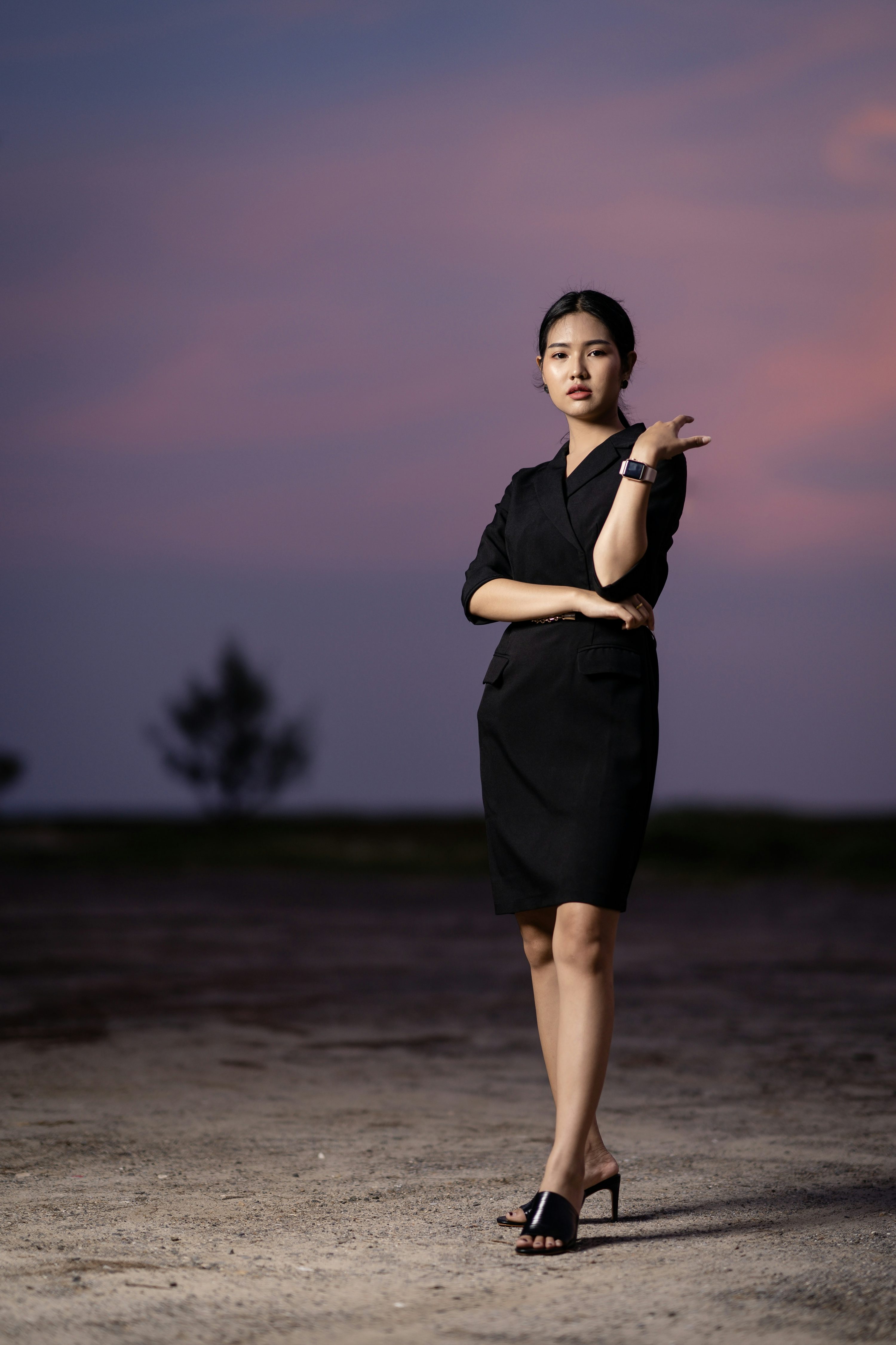 a full shot of a young, slender asian woman in a black dress standing outdoors at sunset. she's posed confidently, one hand on her hip and the other gesturing slightly. the sky is a blend of purple and pink hues, and the ground is a dusty, uneven surface. she's wearing black heeled sandals. the overall mood is sophisticated and stylish.
