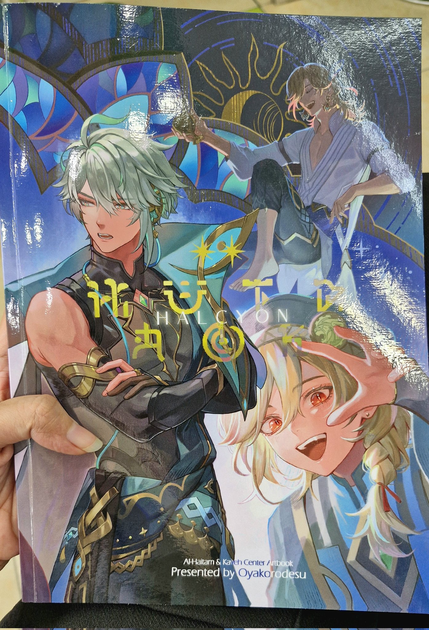 the image shows an artbook cover for genshin impact characters alhaitham and kaveh. the book is titled "halcyon days" and features the two characters in a romantic pose. the cover art is drawn in a colorful anime style.