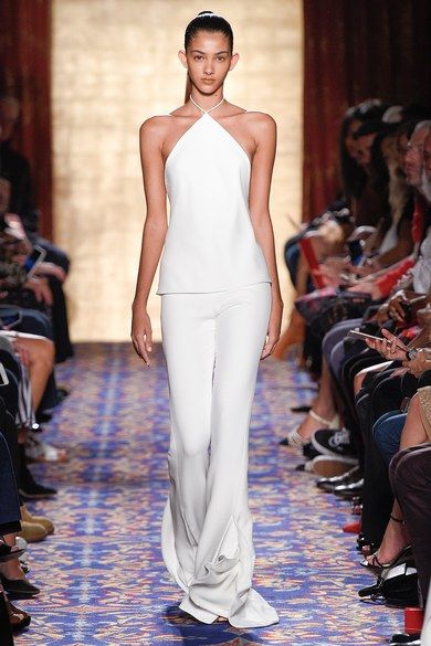 a young woman model walks the runway in a white halter top dress and matching pants. she has dark hair pulled back in a high ponytail, and her makeup is natural. the model is walking confidently and smiling slightly. the audience is seated on both sides of the runway, watching the show. the carpet is a patterned blue and white. the overall style is modern and elegant. the woman's body language is confident and powerful, as she walks down the runway.  the white outfit accentuates her slim build and long legs, highlighting her feminine form. she looks like she is enjoying the experience and taking it in stride.