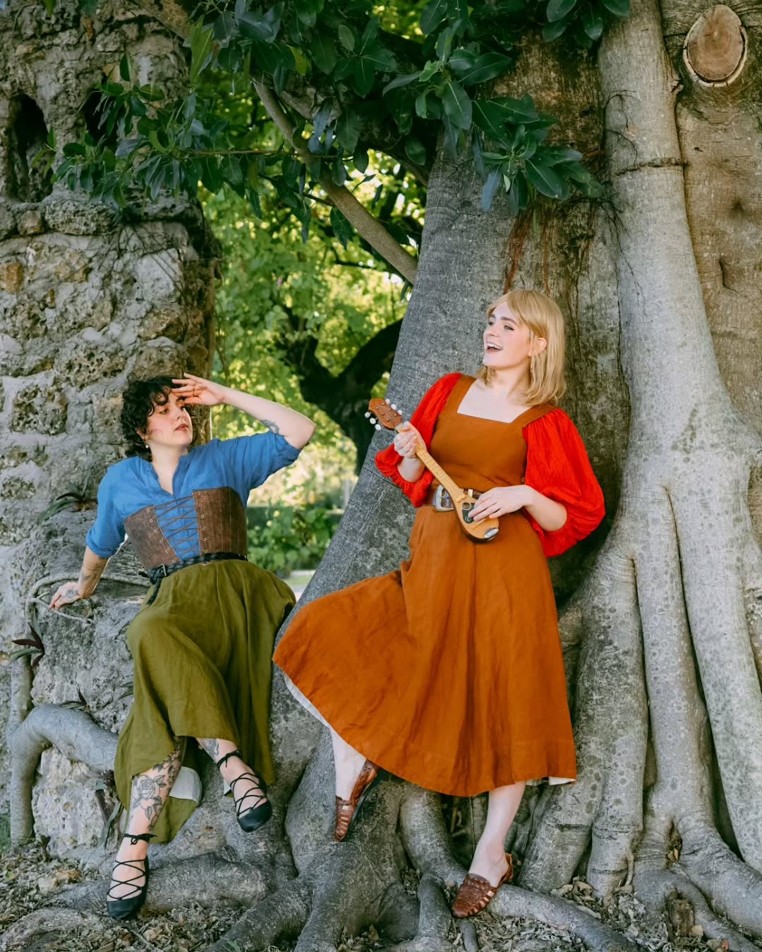 the image shows two women outdoors, positioned against the massive trunk and roots of a large tree. one woman is seated, wearing a blue shirt over a brown corset and a long olivegreen skirt. she has tattoos visible on her legs. the other woman is leaning against the tree, wearing a long, rustcolored dress and holding a bouzoukilike musical instrument.  she's smiling and appears to be playing or posing with the instrument. the scene is sunny and suggests a summer day in a parklike setting. the overall aesthetic leans towards vintage or historical, with a slightly whimsical or fairytale feel to the clothing and setting. the women appear relaxed and happy, possibly friends enjoying a moment together.