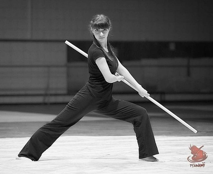 a woman in black pants and a black tshirt performs a martial arts move with a staff. she is in a low stance and is holding the staff with both hands. the image is in black and white.