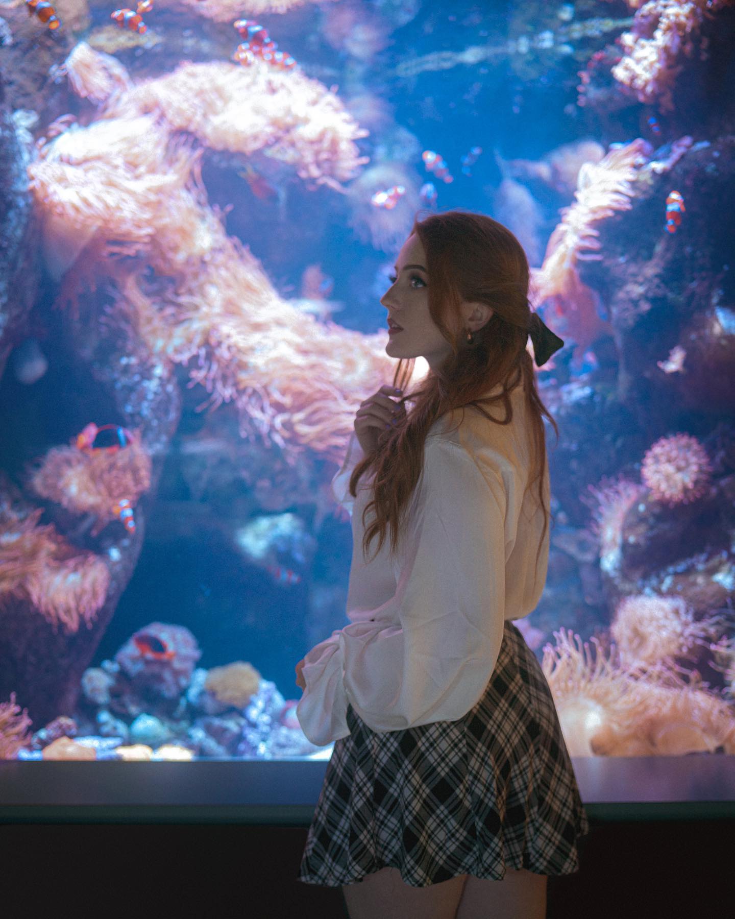 young woman with long red hair, styled with a black bow, stands in profile gazing at an aquarium.  she's wearing a white peasant blouse and a black and white pleated skirt. the aquarium is filled with vibrant coral reefs and various fish. the lighting is primarily artificial, illuminating the scene with a cool blue tone.