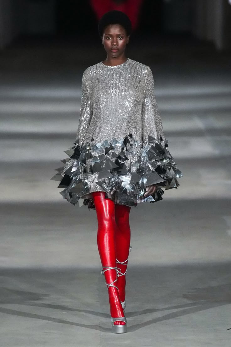 a fashion model walks the runway during a show. she is wearing a silver sequined dress with geometric details at the hem, red tights, and silver platform shoes with wraparound straps.