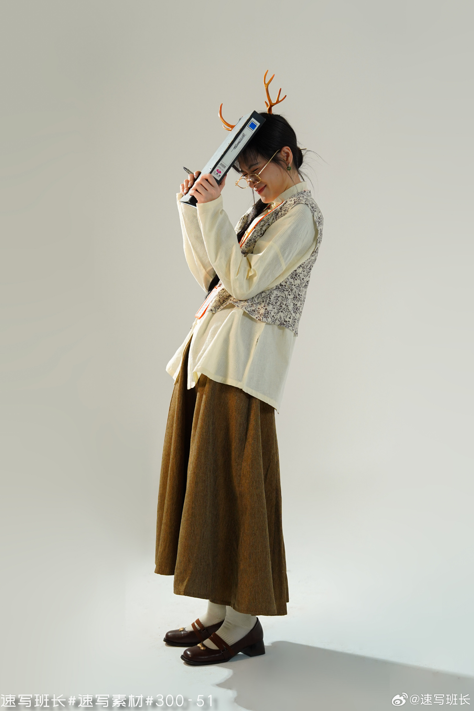 a young woman in a whimsical outfit stands sideways, holding a book above her head. she is wearing deer antlers, a creamcolored cardigan, a brown vest, a brown skirt, white socks, and black mary jane shoes. the background is plain white, and the lighting is soft and artificial.