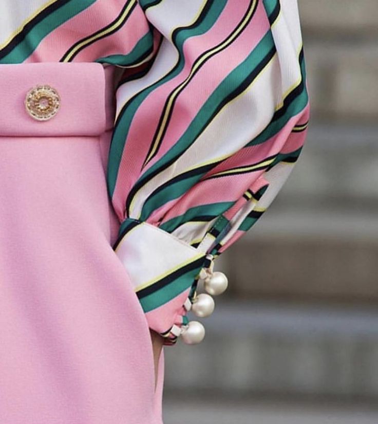 closeup view of a woman's outfit featuring a pink, striped blouse with a unique cuff detail. the cuffs are adorned with a cluster of pearls, adding an elegant touch to the overall design. the blouse is paired with a solid pink skirt, creating a chic and fashionable ensemble. the image showcases the fine details of the fabric and the intricate design elements of the garment.  the overall style is sophisticated and feminine.