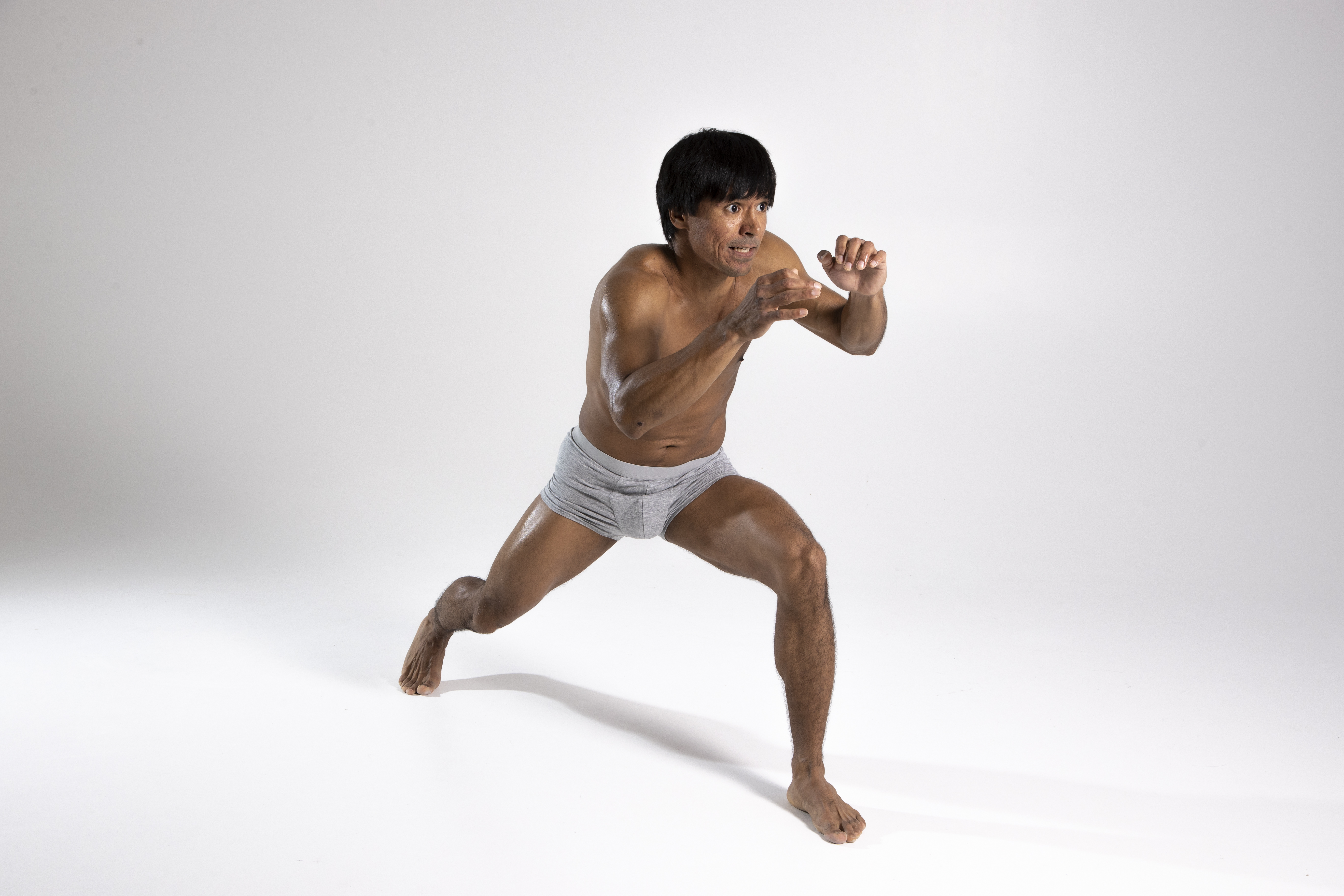 a man in a fighting stance wearing underwear, barefoot, in a studio setting. he is focused, with a lean muscular build and athletic posture.