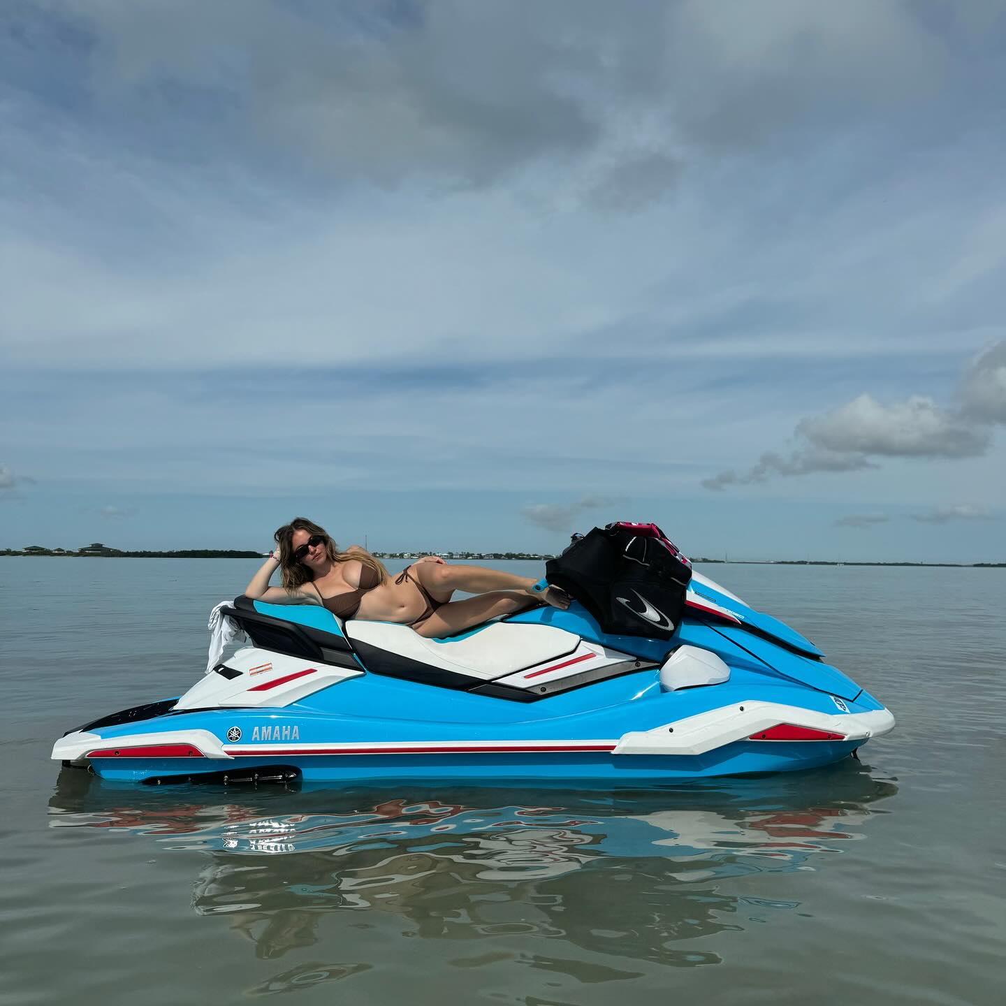 a woman in a brown bikini is lying on a blue and white yamaha jet ski in calm ocean water.  the sky is mostly cloudy but bright.  a black bag is secured to the back of the jet ski. the scene is peaceful and idyllic, suggesting a summer vacation.