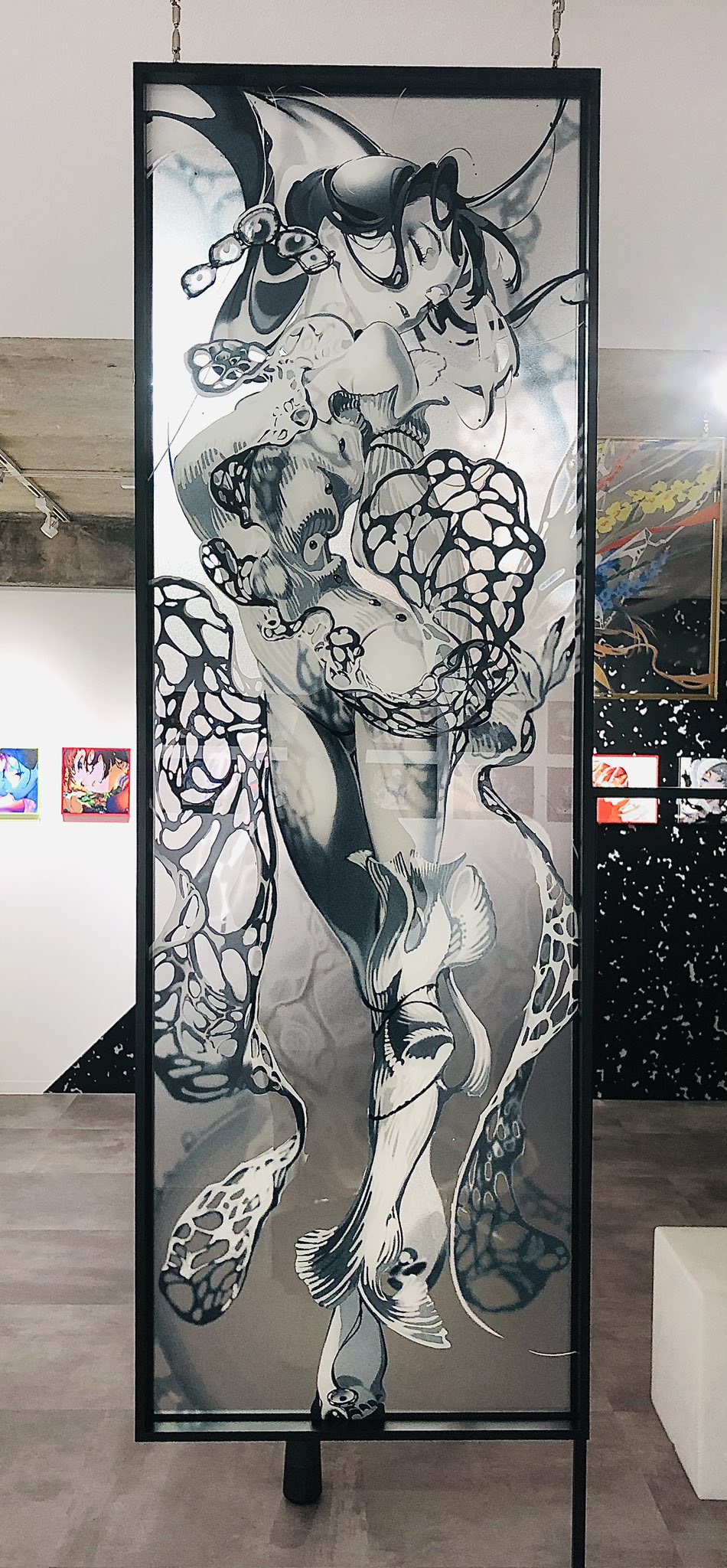 the image showcases a captivating piece of modern art. it's a tall, rectangular artwork made on what appears to be glass or a transparent medium. the art itself features abstract black and white forms resembling a human figure intertwined with organic shapes and patterns. this piece is displayed within an art gallery setting, as suggested by the neutral walls, polished concrete floor, and the presence of other artworks subtly visible in the background.
