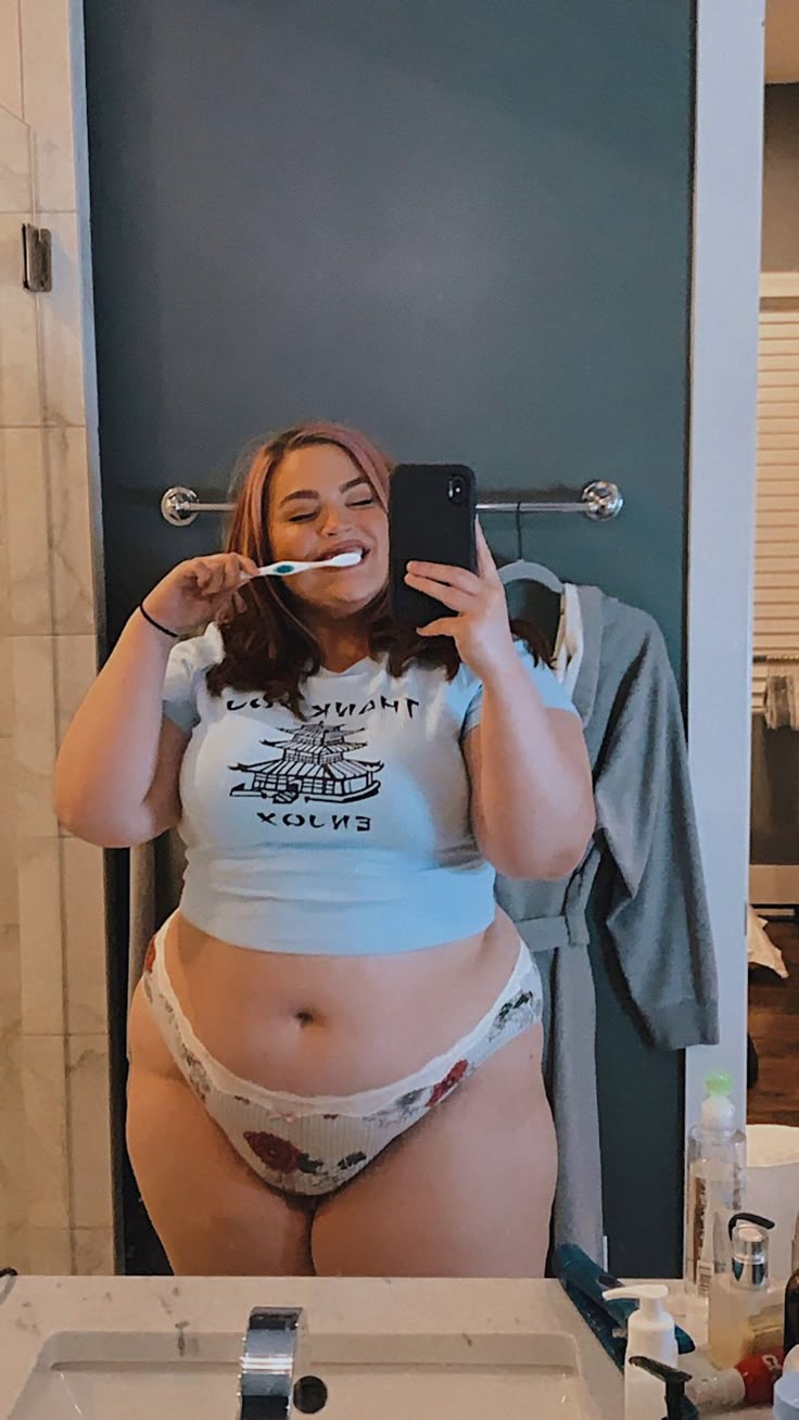 a woman is taking a selfie in a bathroom mirror while brushing her teeth. she is wearing a light blue tshirt and patterned underwear. a gray robe hangs on the wall behind her. there are toiletries on the counter in front of her.