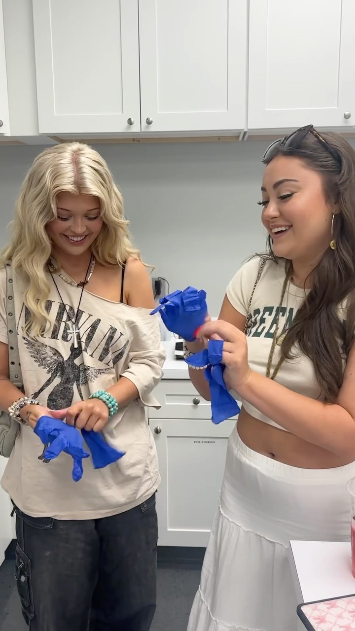 two women are indoors, wearing casual clothing and blue gloves. the woman on the left has blonde wavy hair, wearing a nirvana tshirt, black jeans, and multiple bracelets. the woman on the right has brown hair, wearing a crop top and a long white skirt. they both are smiling and interacting with the gloves. behind them are white cabinets.