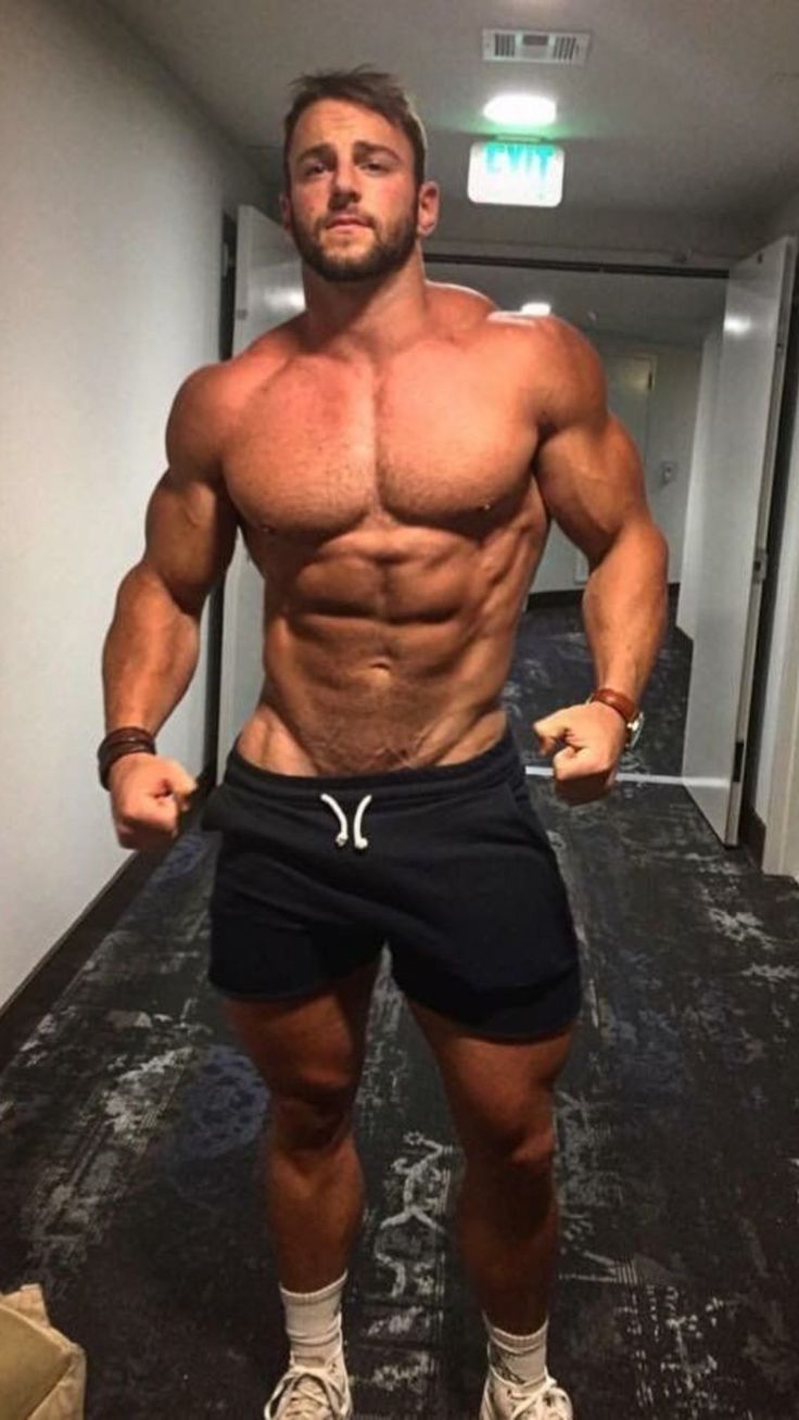 the image shows a muscular man standing in a hallway. he is shirtless and wearing dark shorts, showcasing his welldefined physique. his expression is serious, and he appears to be in good shape. the background is a simple hallway with an exit sign visible.
