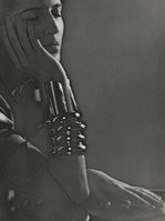 the image is a striking black and white portrait of a woman.  she's captured from the chest up, her face partially obscured by her hand, which rests gently against her cheek.  she appears pensive or thoughtful. her gaze is downcast, adding to the overall mood of introspection. the woman is adorned with elaborate, dark bracelets or cuffs on her wrists, which are prominent features in the image. the lighting is dramatic, using chiaroscuro to emphasize the contours of her face and body. the background is dark and indistinct, drawing attention entirely to the woman. the overall style suggests a vintage aesthetic, possibly from the art deco era. the photograph has a high artistic quality, demonstrating skillful use of light, shadow, and composition.