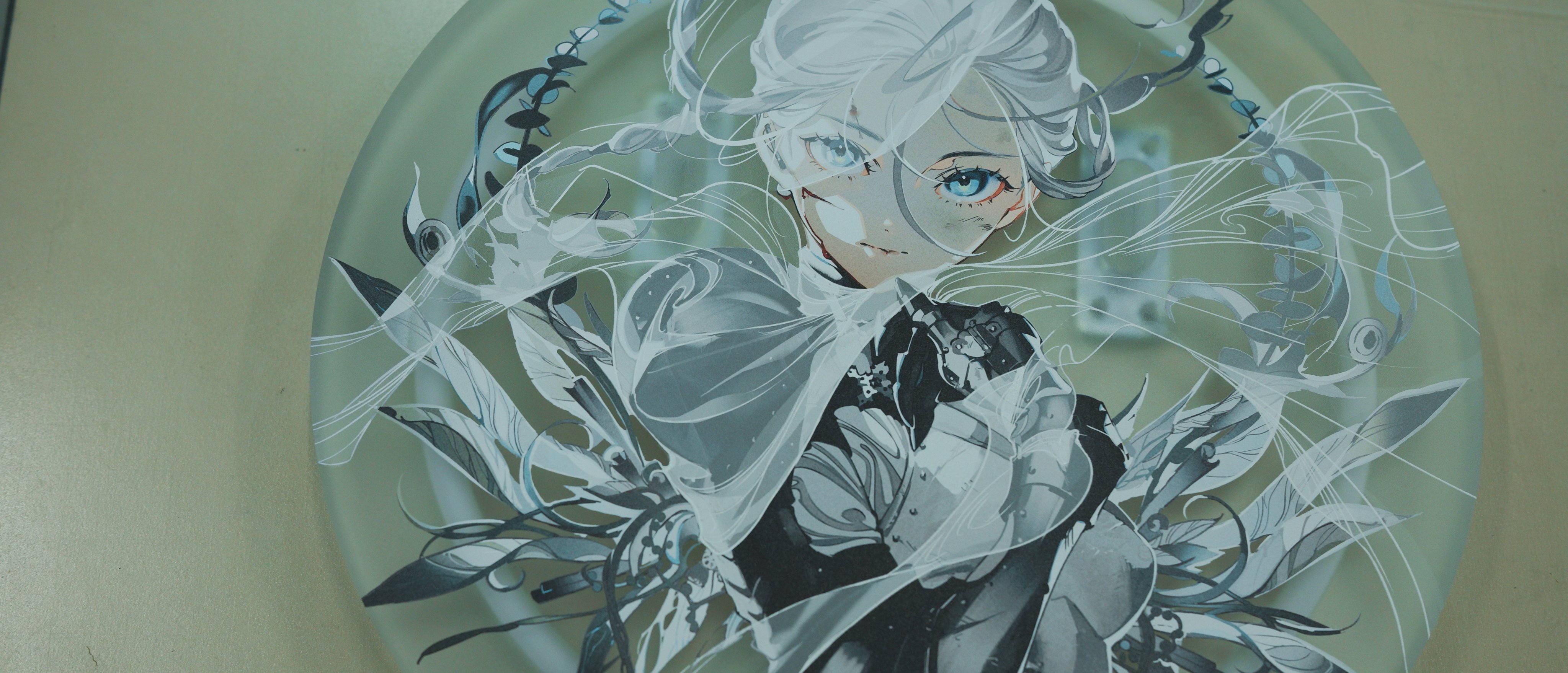 this is a top down view of a circular glass artwork featuring a black and white illustration of an anime girl. her white hair appears to be flowing in the wind. there are blue and white accents in the artwork.