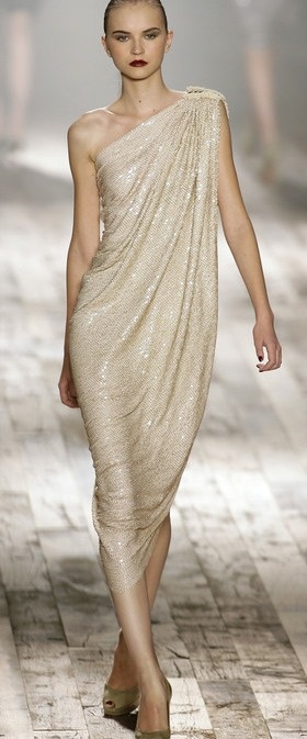 a fashion model walks the runway in a light beige one shoulder sequined dress.  she is posing with her left hand on her hip and right hand extended to the side. the backdrop is a light wood runway with a white wall in the background.