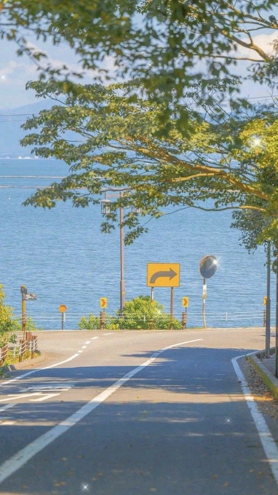 the image shows a road curving to the right toward a body of water. there are trees on both sides of the road, and a blue sky with some clouds is visible in the background. the photo gives off a sense of peace and tranquility.