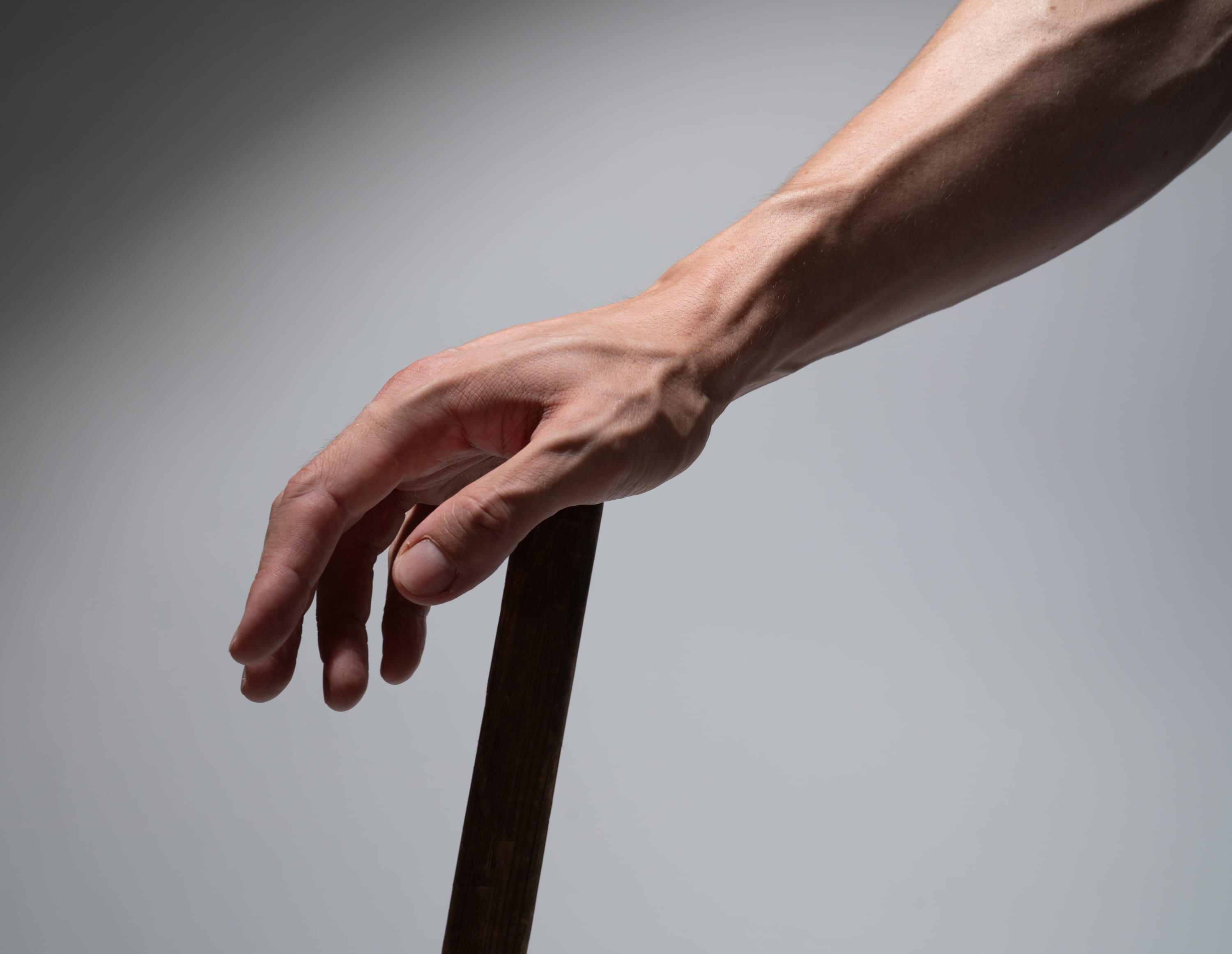 this image shows a man's muscular arm reaching down towards the left side of the frame to grasp a dark brown wooden pole. the arm is illuminated by bright studio lighting, creating strong contrasts and highlighting the contours of the muscles and tendons. the background is a smooth, light gray, providing a minimalist setting that emphasizes the subject.