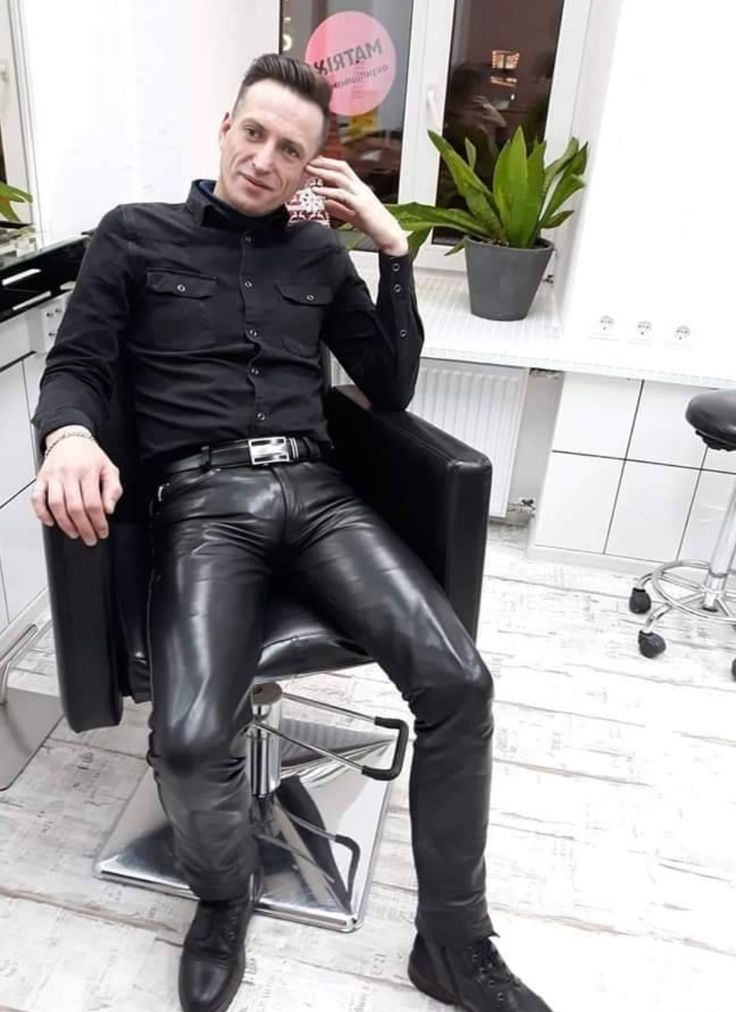 a stylish man with short hair sits in a barber chair, posing for a photo. he is wearing a black buttondown shirt, black leather pants, and black boots. the setting appears to be a hair salon, with a plant visible in the background. the overall impression is one of fashion and cool style.