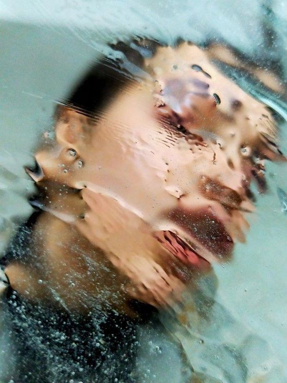 the image is a closeup, abstract portrait of a person's face viewed through a layer of water or condensation on a glass surface. the water distorts the features, making them blurry and indistinct. only parts of the face are visible, including eyes and mouth. the overall effect is ethereal and dreamlike. the colors are muted and pastel, with a predominance of blues and greens. the water droplets and textures add to the abstract feel of the image.  the mood is somewhat melancholic and mysterious.