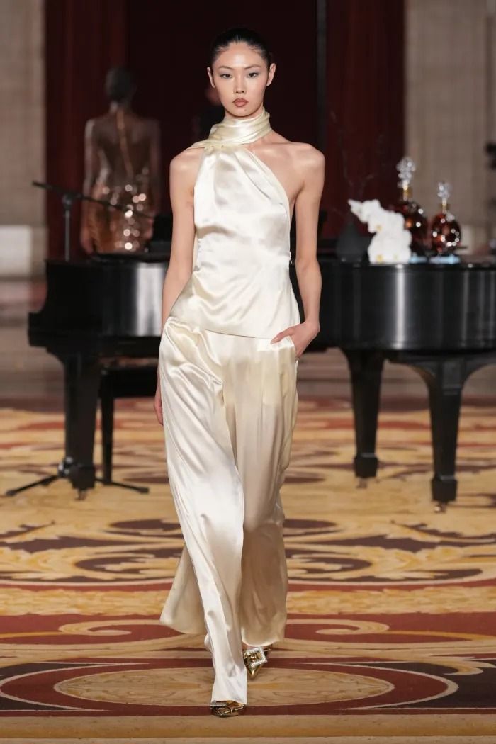 a model walks the runway during a fashion show. she is wearing a oneshoulder silk top and wideleg pants in a cream color. in the background there's a piano with a few bottles of cognac. the runway is covered with a patterned carpet.