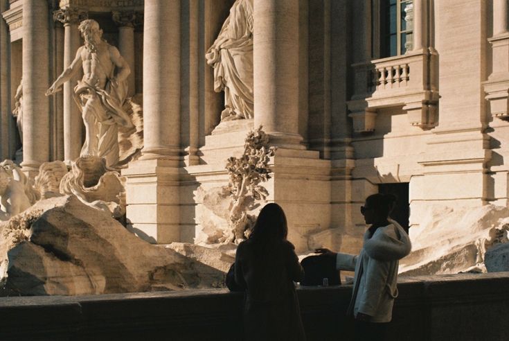 two women stand silhouetted against the backdrop of the trevi fountain in rome.  the iconic baroque fountain is partially visible, showcasing its intricate sculptures and classical architecture. the warm sunlight casts shadows on the stonework, highlighting the textures and details of the building.  the scene evokes a sense of history, art, and the beauty of roman architecture.