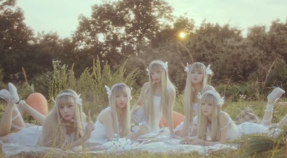 five women are lying on their stomachs on a blanket spread out in a field. the women are wearing white dresses with feathery white wings on their heads. they have a picnic basket in front of them and trees in the background. the lighting is soft and golden, suggesting the time of day is sunset.