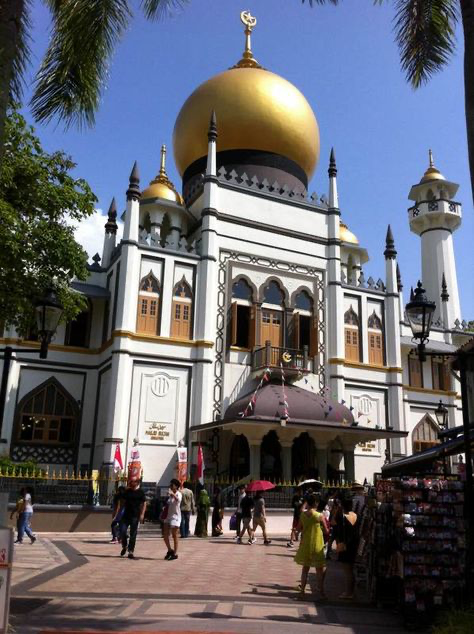 a vibrant photo showcasing the masjid sultan in singapore on a sunny day. the mosque's golden dome and intricate architecture are prominent, surrounded by lush palm trees and bustling activity. tourists and locals are visible, with shops selling souvenirs adding to the scene's lively atmosphere. the image offers a mix of architectural beauty and everyday urban life, capturing the essence of singapore's multicultural heritage.