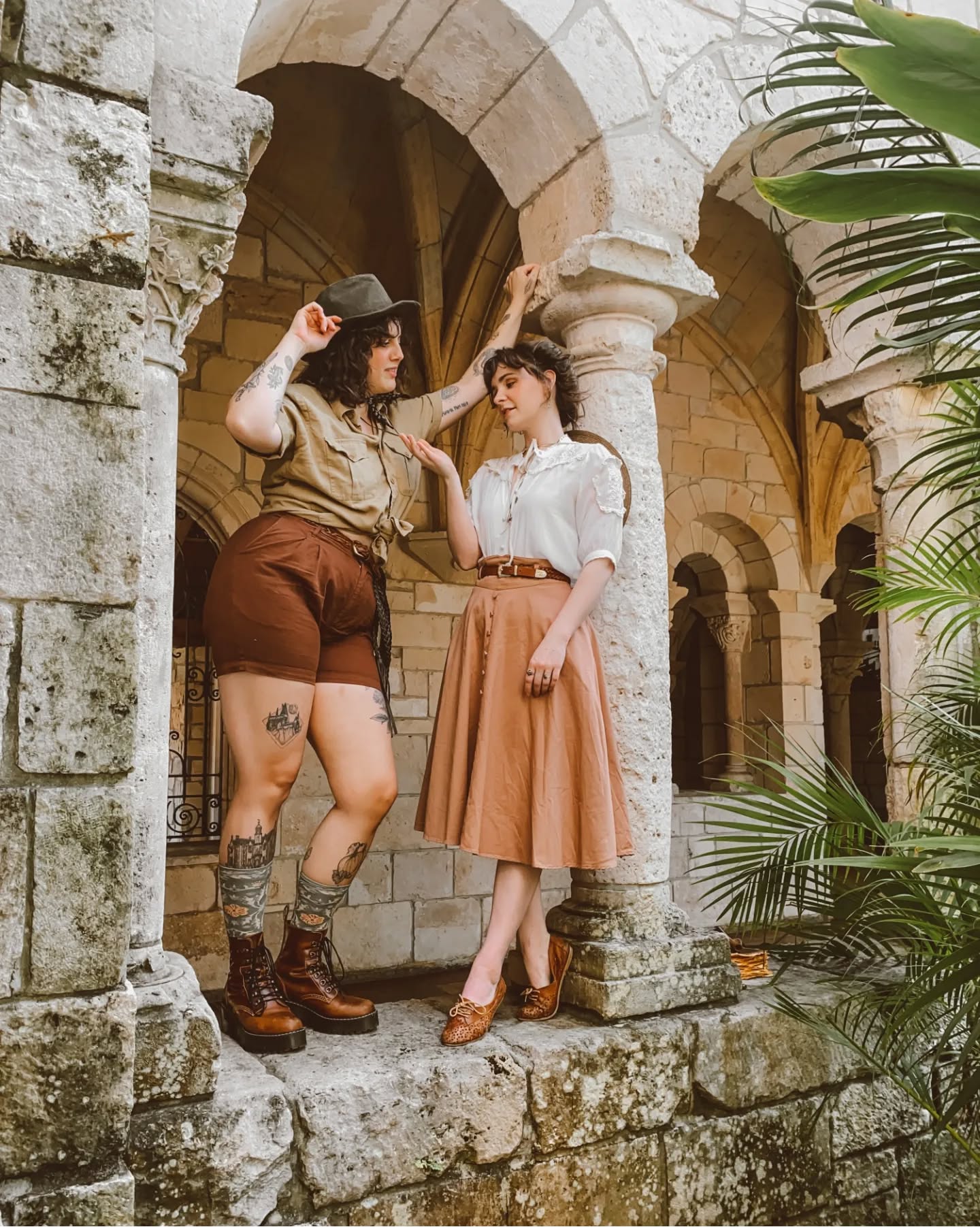 the image shows two women dressed in vintageinspired clothing posing in a stone cloister. one woman is wearing khaki shorts and a matching shirt with a hat, while the other woman wears a light brown skirt and a white blouse. both are accessorized with brown shoes. they are positioned under a stone archway with lush tropical foliage in the background. the style is reminiscent of 1930s or 1940s fashion, creating a retro aesthetic. the overall mood is one of adventure and exploration. the background shows a richly detailed historical stone building.