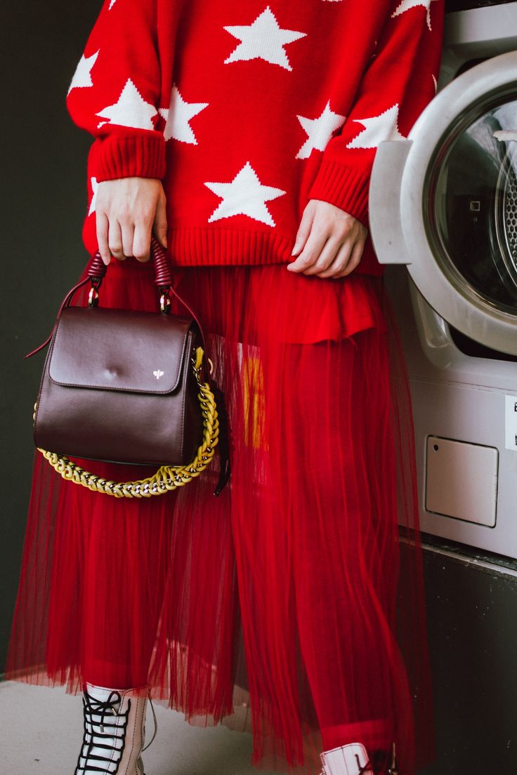 a woman is standing next to a washing machine. she is wearing a red sweater with white stars, a long red tulle skirt, burgundy handbag with a gold chain strap and white boots. the overall style is fashionable and chic.