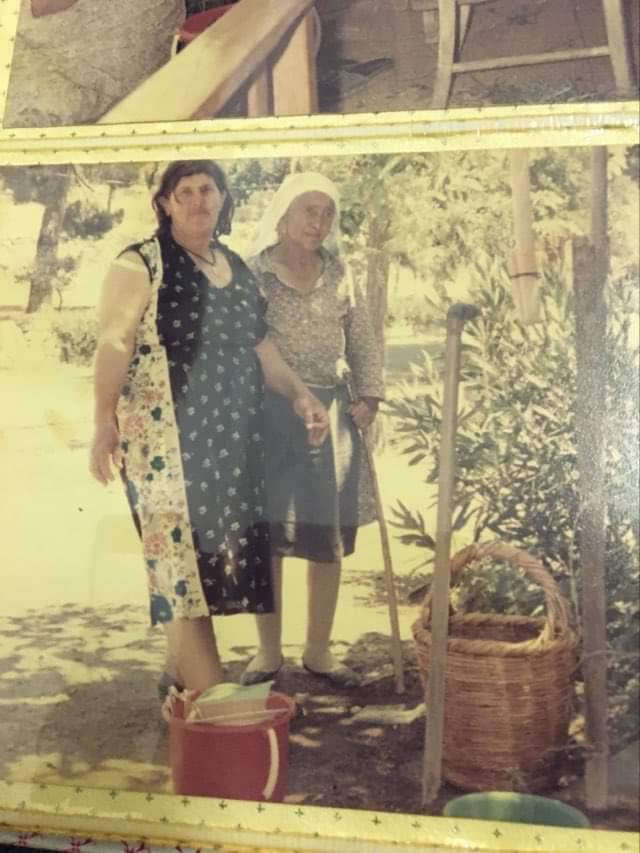 he image is an old photograph showing two women standing outdoors in a gardenlike setting. the woman on the left is younger, wearing a floral patterned dress and apron. the woman on the right is older, wearing a headscarf and a darker dress. they are standing near a wicker basket and a bucket. both women appear to be of mediterranean descent. the background suggests a rural or countryside setting. the photo has a vintage feel, with faded colors and slightly blurred details.