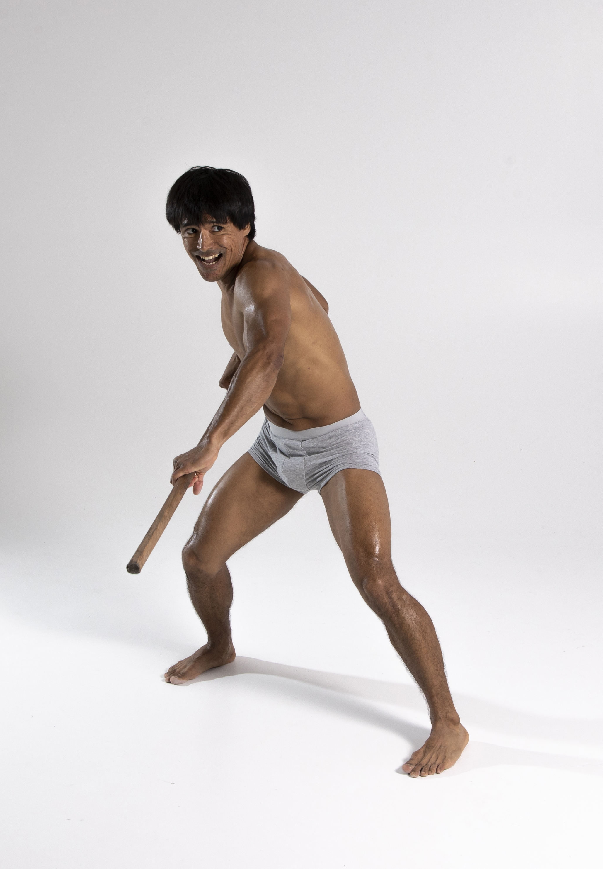 a shirtless man wearing underwear is holding a stick while smiling and looking at the camera. he is barefoot and appears muscular. the background is a plain white studio backdrop.