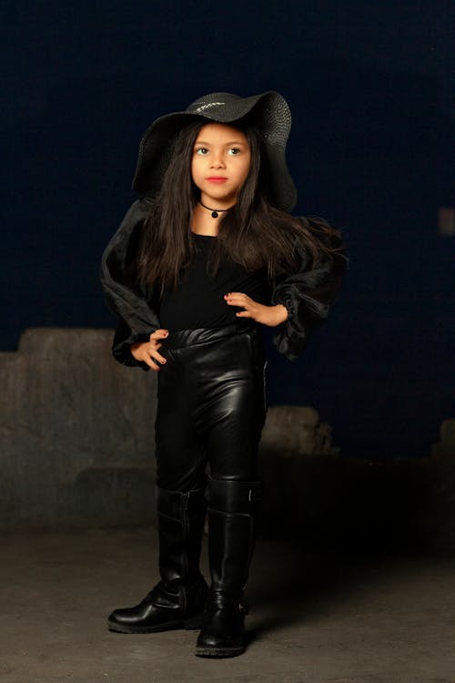 a young girl, possibly a child model, is striking a pose in a studio setting. she is dressed in a stylish allblack outfit consisting of a black hat, a black top, black leather pants, and tall black boots.  her pose is confident and slightly defiant. the background is dark and moody, enhancing the overall dramatic effect of the image. the girl's expression is serious. the overall aesthetic is edgy and fashionable.