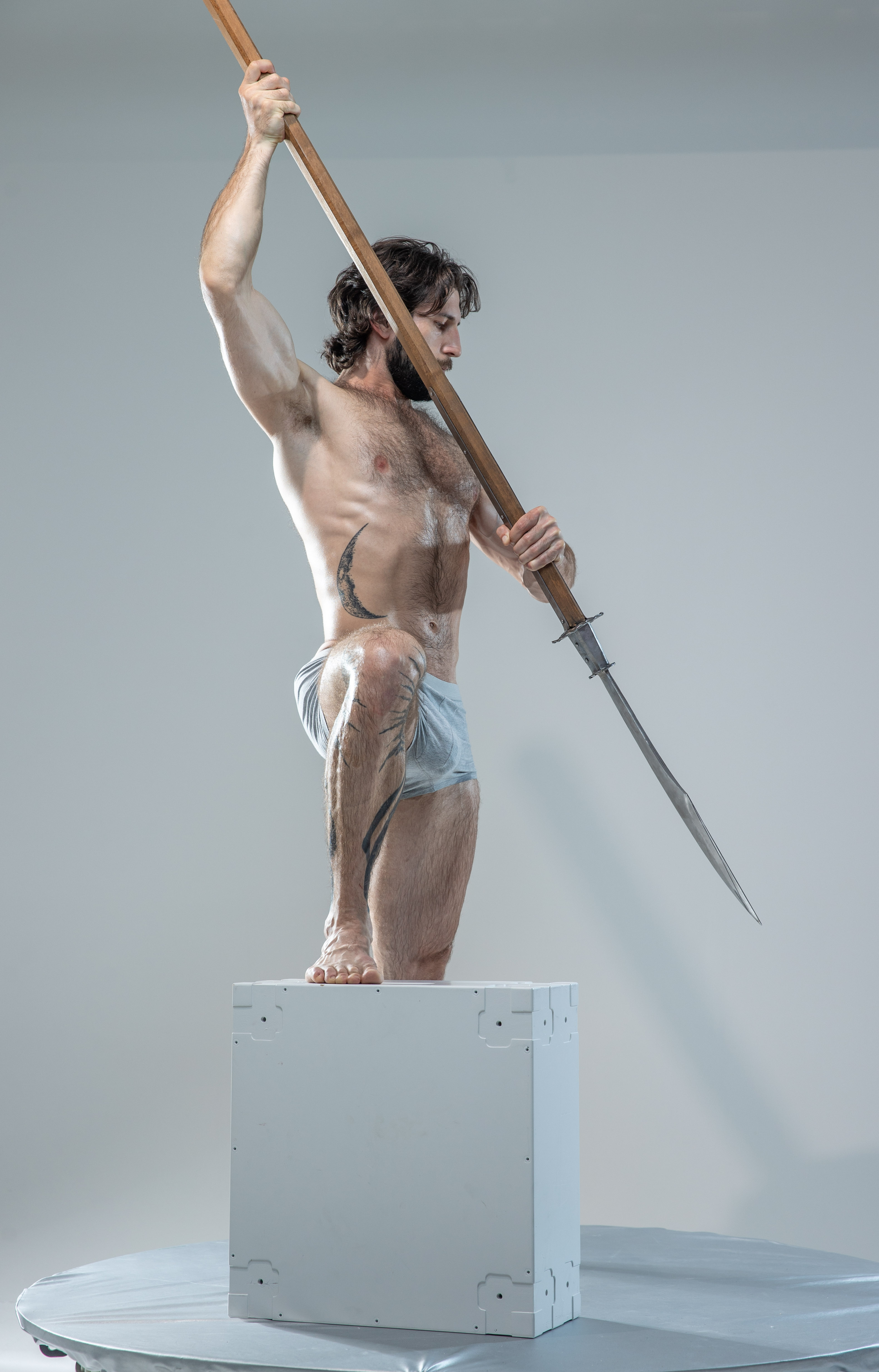 a shirtless, muscular man with a beard and tattoos poses dynamically on a white cube in a photography studio. he holds a halberd aloft with both hands, gazing intently towards the spearhead. the background is a plain, light gray, and the lighting is soft and even, highlighting the man's physique.