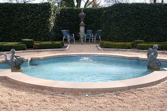 this image shows a formal garden with a circular fountain as the main focal point. the fountain has two statues on either side, one of a woman riding a sea creature, and another of two children on a sea creature. the fountain is surrounded by gravel. in the background, there is a seating area with four blue chairs surrounding a tall urn, all within a space created by tall, manicured hedges. there are also neatly trimmed box hedges near the fountain.