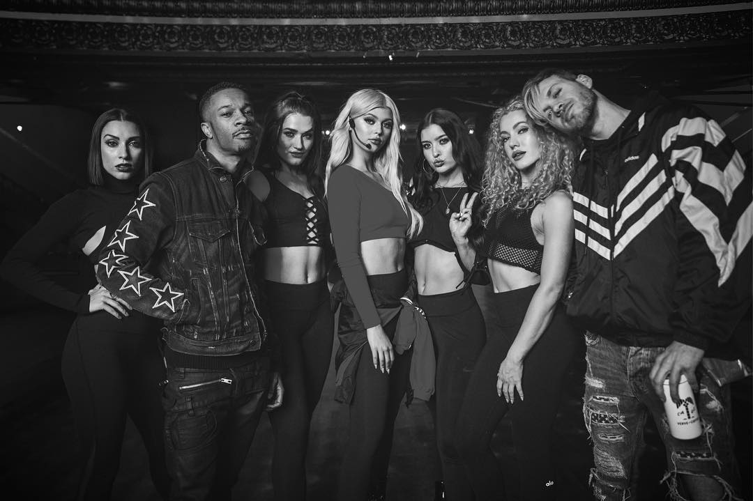 a black and white group photo of six women and two men posing together. the people are all stylishly dressed, with the women wearing crop tops, bodysuits, and sportswear, and the men wearing a denim jacket with stars, and a track jacket with ripped jeans. one of the women is wearing a microphone headset.