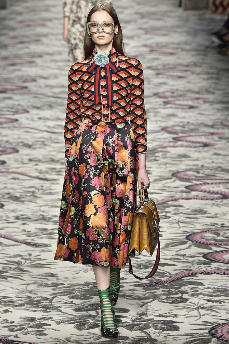 a model walks the runway at a gucci fashion show. she is wearing a geometric patterned blouse and a floral midi skirt, paired with green laceup sandals and carrying a gold handbag.  the runway features a patterned floor. the overall style is high fashion and sophisticated.